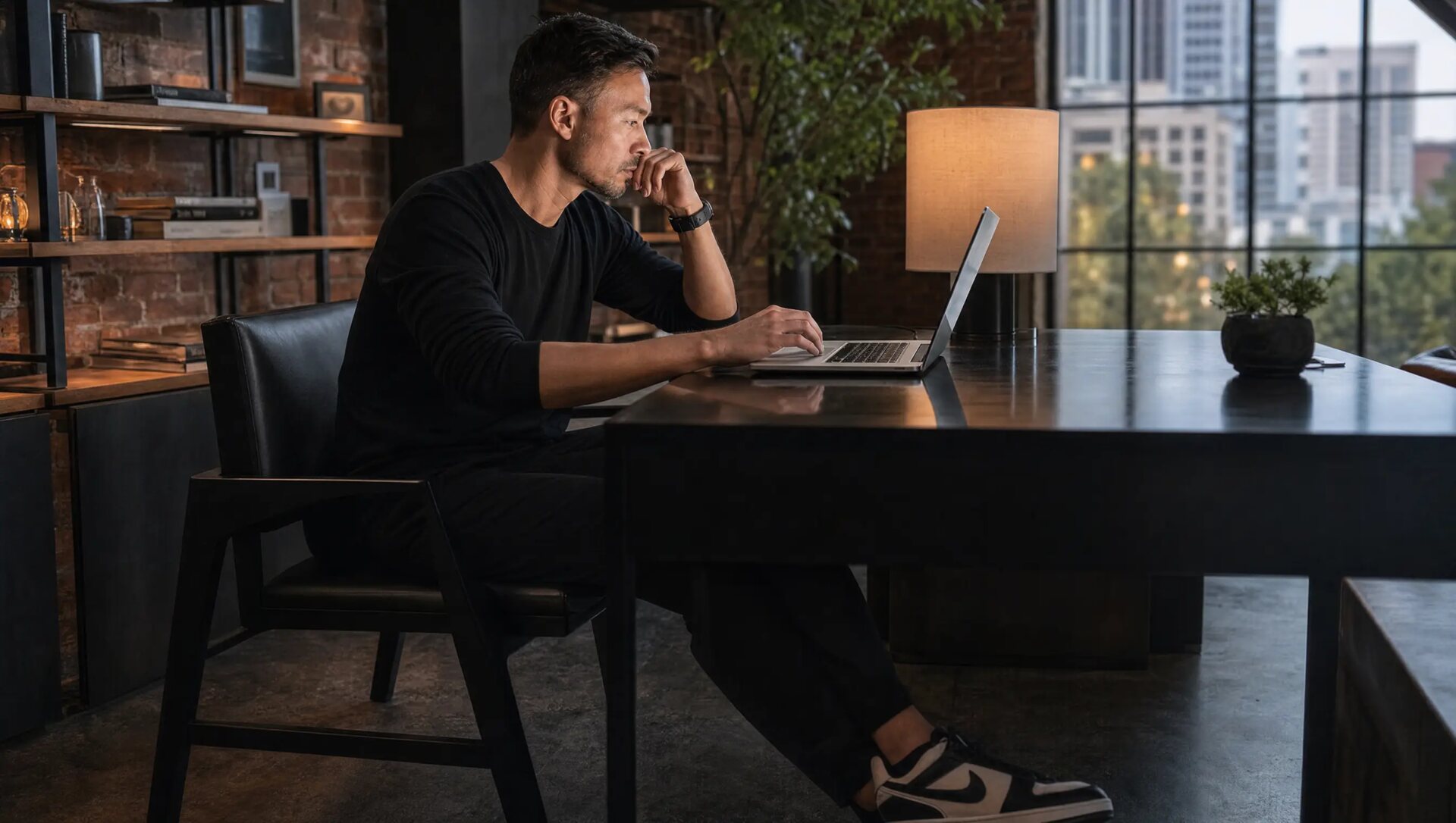 A designer working at a laptop in a brick loft studio at dusk, a lamp lit beside the window.