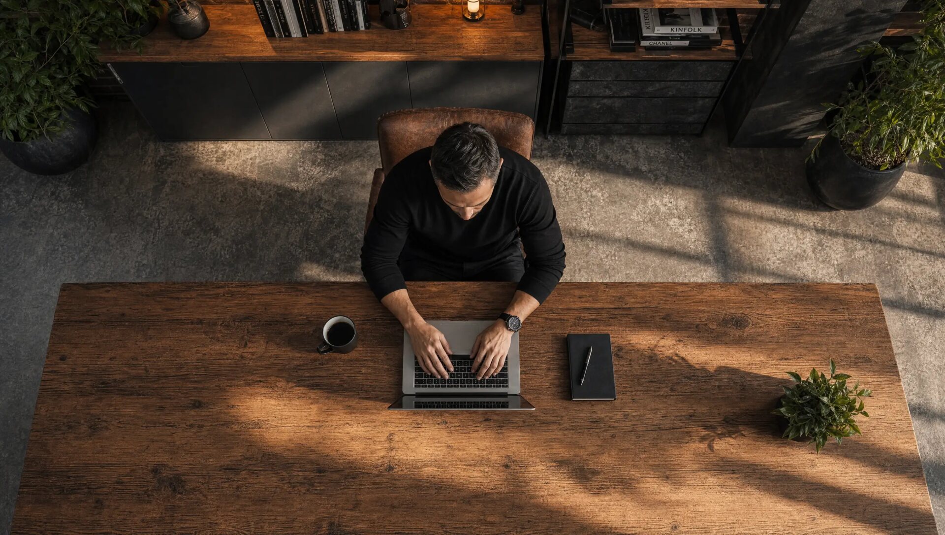 Seen from above: a man at a laptop at a long wooden desk in a sunlit loft, a coffee and a notebook beside him.